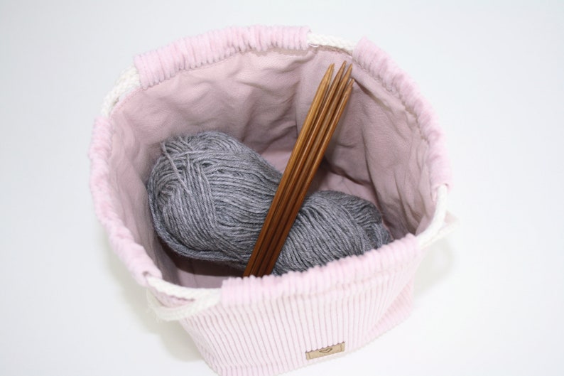 Puede incluir: Una bolsa de cord&oacute;n de pana rosa con forro blanco. La bolsa est&aacute; llena de dos ovillos de hilo gris y un juego de agujas de tejer de madera.