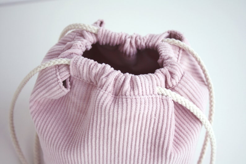 Puede incluir: Una bolsa de cord&oacute;n de pana rosa con cordones blancos. La bolsa est&aacute; hecha de una tela suave y texturizada y tiene una abertura ancha.