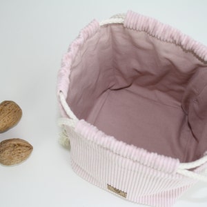 Puede incluir: Una bolsa de cord&oacute;n de pana rosa con cordones blancos. La bolsa est&aacute; vac&iacute;a y tiene un forro rosa claro. Hay tres nueces en primer plano.