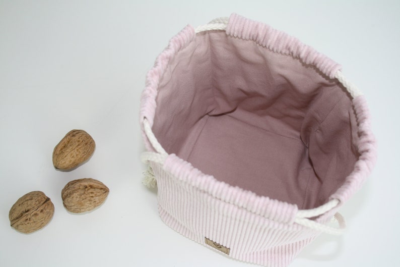 Puede incluir: Una bolsa de cord&oacute;n de pana rosa con cordones blancos. La bolsa est&aacute; vac&iacute;a y tiene un forro rosa claro. Hay tres nueces en primer plano.