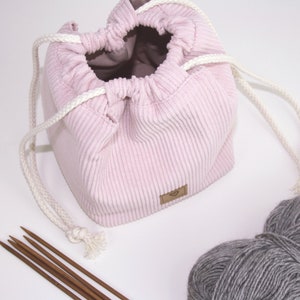 Puede incluir: Una bolsa de cord&oacute;n de pana rosa con cordones blancos, una peque&ntilde;a etiqueta marr&oacute;n y una bola de hilo gris a su lado.
