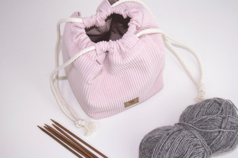 Puede incluir: Una bolsa de cord&oacute;n de pana rosa con cordones blancos, una peque&ntilde;a etiqueta marr&oacute;n y una bola de hilo gris a su lado.