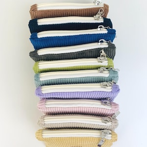 May include: A stack of nine zippered pouches in various colors, including brown, blue, gray, green, light blue, purple, pink, beige, and yellow. The pouches are made of a corduroy fabric and have a white zipper.