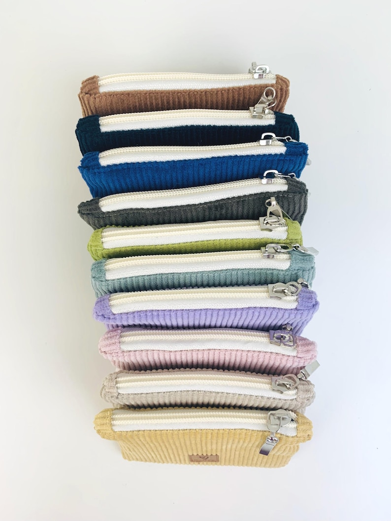 May include: A stack of nine zippered pouches in various colors, including brown, blue, gray, green, light blue, purple, pink, beige, and yellow. The pouches are made of a corduroy fabric and have a white zipper.
