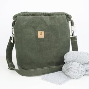 Könnte beinhalten: Eine grüne Cord-Kordeltasche mit einem langen Schultergurt und einem kleinen Lederpatch mit einem Herzlogo, auf dem "MYLOVE" steht. Die Tasche liegt auf einer weißen Oberfläche mit zwei Knäuelen grauer Wolle und einem grauen Häkelquadrat.