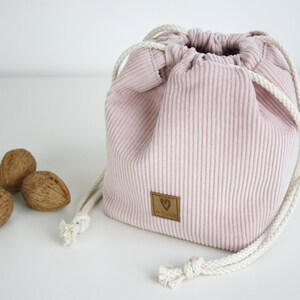 Puede incluir: Una bolsa de cord&oacute;n de pana rosa con cordones blancos y una etiqueta de cuero marr&oacute;n con un coraz&oacute;n y el texto "De Kleine" en ella. Tres nueces est&aacute;n en primer plano.