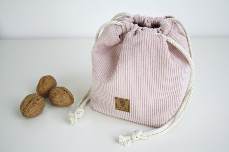 Puede incluir: Una bolsa de cord&oacute;n de pana rosa con cordones blancos y una etiqueta de cuero marr&oacute;n con un coraz&oacute;n y el texto "De Kleine" en ella. Tres nueces est&aacute;n en primer plano.