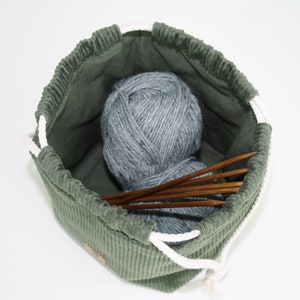 May include: A green corduroy drawstring bag with a white drawstring, containing a ball of grey yarn and wooden knitting needles.