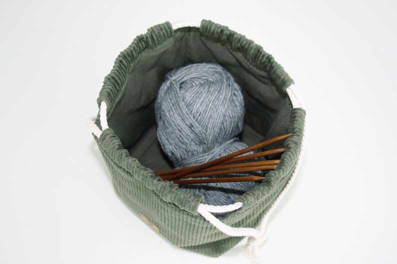 May include: A green corduroy drawstring bag with a white drawstring, containing a ball of grey yarn and wooden knitting needles.