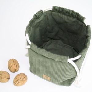 May include: A green corduroy drawstring bag with white cords. The bag is empty and sitting on a white surface. Three walnuts are in front of the bag.