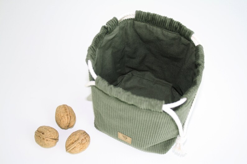 May include: A green corduroy drawstring bag with white cords. The bag is empty and sitting on a white surface. Three walnuts are in front of the bag.