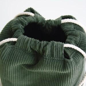 May include: A green corduroy drawstring bag with white cords. The bag is empty and the cords are gathered at the top.