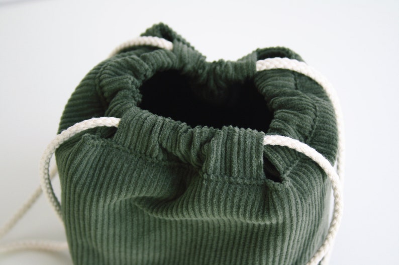 May include: A green corduroy drawstring bag with white cords. The bag is empty and the cords are gathered at the top.