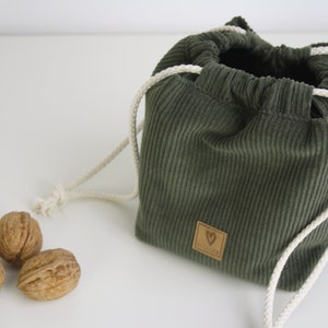 May include: A green corduroy drawstring bag with white cords and a brown leather heart-shaped tag. The bag is sitting on a white surface with three walnuts in front of it.