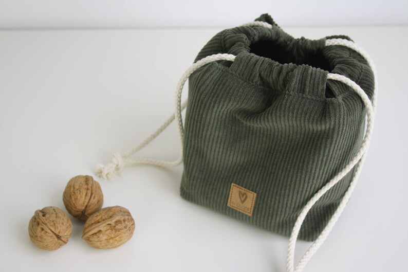 May include: A green corduroy drawstring bag with white cords and a brown leather heart-shaped tag. The bag is sitting on a white surface with three walnuts in front of it.