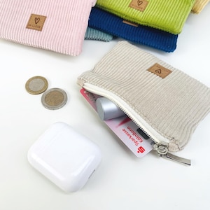May include: A beige corduroy zippered pouch with a small heart-shaped logo. The pouch is open and contains a credit card, a small silver object, and coins.