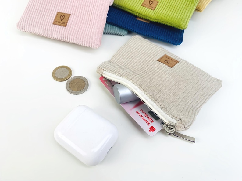 May include: A beige corduroy zippered pouch with a small heart-shaped logo. The pouch is open and contains a credit card, a small silver object, and coins.