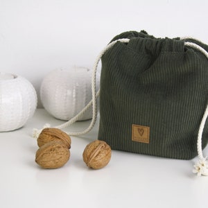 May include: A green corduroy drawstring bag with a brown leather label that says "V&Auml;CK&Ouml;S&Ouml;K". The bag is sitting on a white surface with three walnuts.