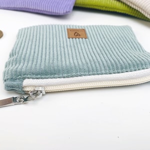 May include: A small, light blue corduroy zipper pouch with a white zipper and a brown leather tag with a heart symbol. The pouch is lying on a white surface.