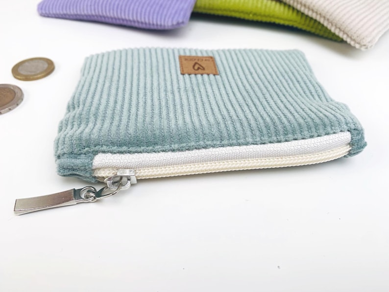 May include: A small, light blue corduroy zipper pouch with a white zipper and a brown leather tag with a heart symbol. The pouch is lying on a white surface.