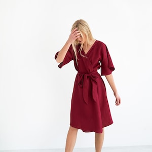 May include: A burgundy wrap dress with a V-neck and a matching waist tie. The dress has short sleeves and falls to the mid-calf. The model is wearing black slip-on shoes.