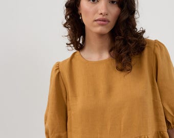 Könnte beinhalten: Senffarbenes Leinenkleid mit kurzen, gepufften Ärmeln und geraffter Taille. Das Kleid hat einen runden Ausschnitt und eine lockere Passform. Der Stoff scheint leicht und atmungsaktiv zu sein, geeignet für wärmeres Wetter. Das Model trägt kleine goldene Ohrringe.