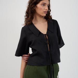 K&ouml;nnte beinhalten: Schwarzes kurz&auml;rmeliges Oberteil mit Kragen und Bindeband. Das Oberteil wird mit einer hoch taillierten, olivgr&uuml;nen Hose getragen. Das Outfit besteht aus einem leichten Stoff, und das Oberteil hat einen V-Ausschnitt. Das Model tr&auml;gt das Outfit in einem Studio.