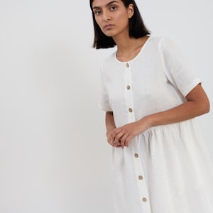 May include: A white linen dress with short sleeves and a button-down front. The dress features a round neckline and a gathered waist. The buttons are a light brown color. The model is wearing the dress.
