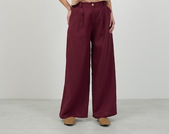 May include: A woman is wearing a light brown sleeveless top and wide-legged burgundy linen pants. She is standing on a white background.