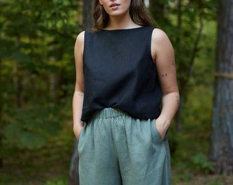 May include: A woman wearing a black sleeveless top and green linen pants stands in a forest setting.