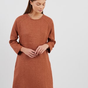 May include: A rust-colored dress with long sleeves and a round neckline. The dress has a simple, classic design with a subtle texture. The model is wearing the dress, which falls to the knees.