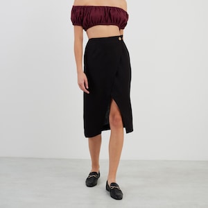 May include: A woman is wearing a burgundy off-the-shoulder crop top and a black wrap skirt with a gold button. She is wearing black loafers with gold buckles.