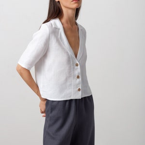 May include: A white linen shirt with a collar and button closure. The shirt has a relaxed fit and is cropped at the waist. The model is wearing the shirt with dark gray pants.