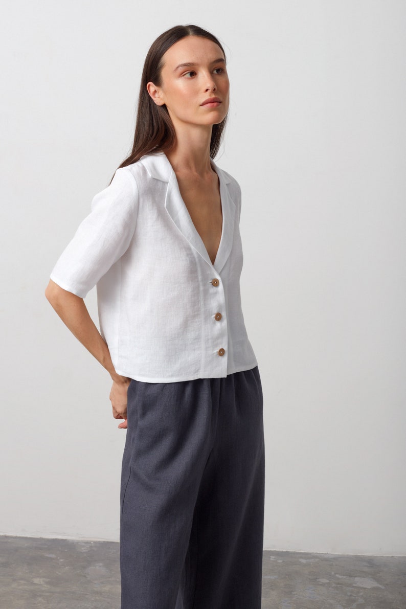 May include: A white linen shirt with a collar and button closure. The shirt has a relaxed fit and is cropped at the waist. The model is wearing the shirt with dark gray pants.