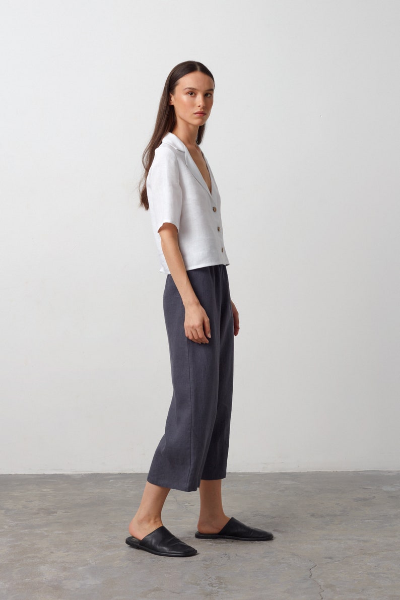 May include: A woman wearing a white linen shirt with a collar and a pair of dark gray linen pants. She is wearing black leather slip-on shoes.