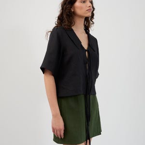 K&ouml;nnte beinhalten: Ein schwarzes kurz&auml;rmeliges Oberteil mit Kragen und Bindeband, kombiniert mit einem gr&uuml;nen Minirock. Das Outfit besteht aus einem leichten Stoff und schafft ein l&auml;ssiges und stilvolles Ensemble. Der Hintergrund ist schlicht wei&szlig;.