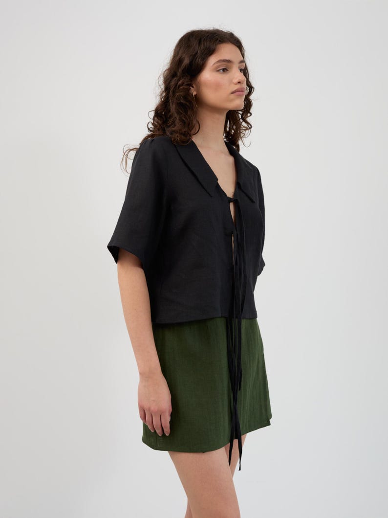 K&ouml;nnte beinhalten: Ein schwarzes kurz&auml;rmeliges Oberteil mit Kragen und Bindeband, kombiniert mit einem gr&uuml;nen Minirock. Das Outfit besteht aus einem leichten Stoff und schafft ein l&auml;ssiges und stilvolles Ensemble. Der Hintergrund ist schlicht wei&szlig;.