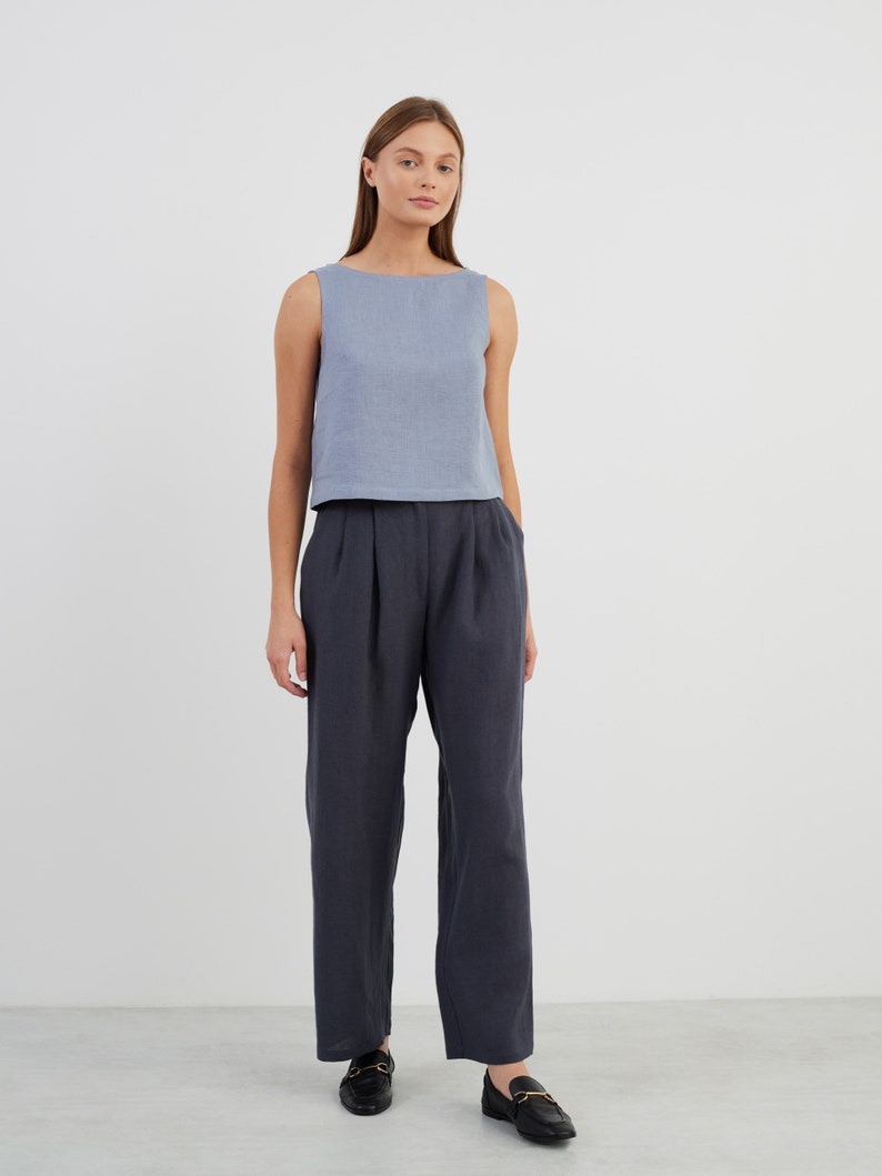 May include: A woman is wearing a light blue sleeveless linen top and dark blue linen trousers. She is wearing black loafers with gold buckles.