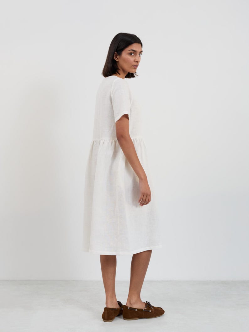 Side-back view of the DOVE linen midi dress in Ivory showing the short sleeve, gathered waist seam, and midi-length skirt