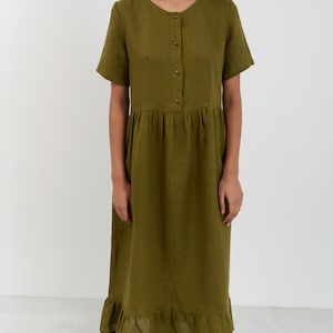 Full-length front view of the SAGE linen midi dress in Moss Green showing round neckline, button-front bodice, gathered drop waist, and ruffle hem with hand in pocket