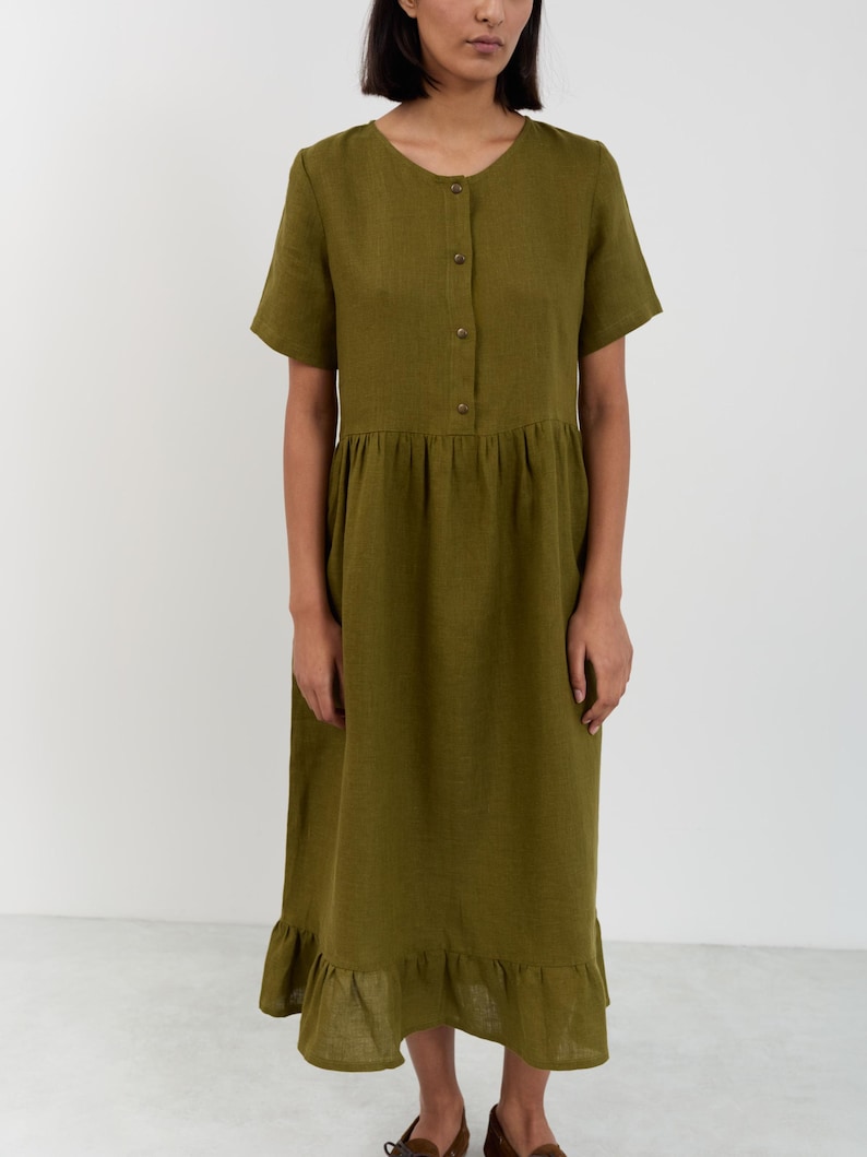 Full-length front view of the SAGE linen midi dress in Moss Green showing round neckline, button-front bodice, gathered drop waist, and ruffle hem with hand in pocket