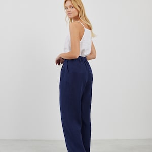 May include: A woman wearing a white tank top and dark blue wide-leg pants. She is standing on a white floor and looking over her shoulder.