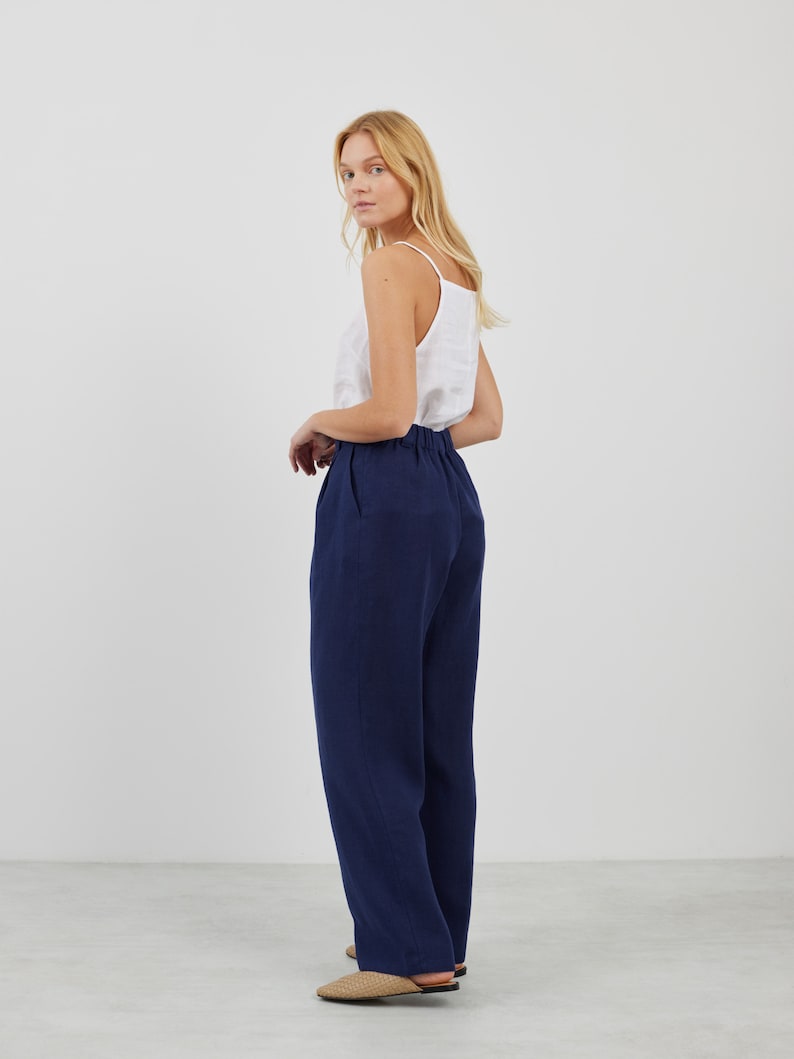 May include: A woman wearing a white tank top and dark blue wide-leg pants. She is standing on a white floor and looking over her shoulder.