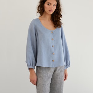 May include: A light blue linen blouse with a scoop neck and button front. The blouse has long sleeves with gathered cuffs and a relaxed fit.