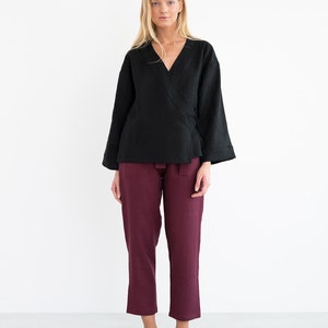 Op de afbeelding: Een zwart linnen wikkeltop met lange mouwen en een V-hals. Het model draagt het top met een bordeauxrode linnen broek en zwarte schoenen.