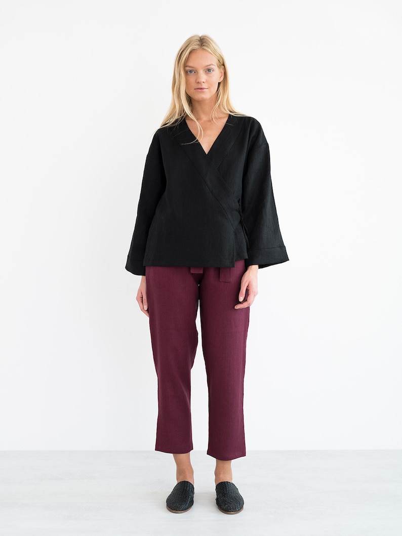 Op de afbeelding: Een zwart linnen wikkeltop met lange mouwen en een V-hals. Het model draagt het top met een bordeauxrode linnen broek en zwarte schoenen.