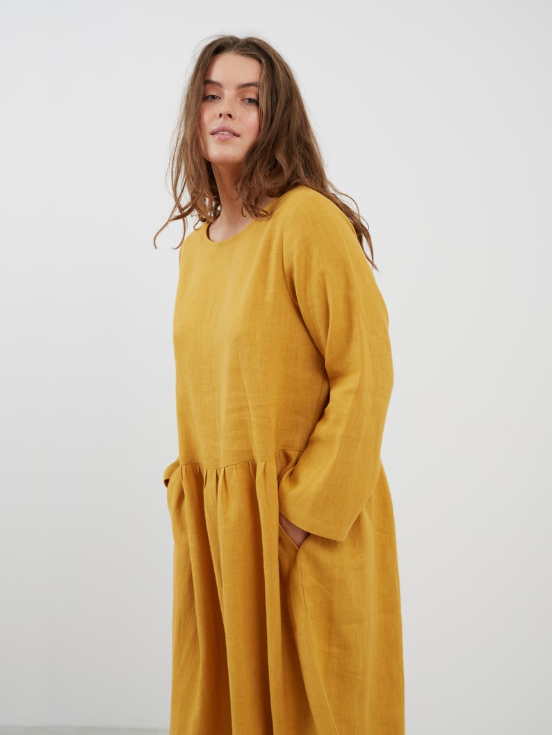 May include: A long-sleeved, yellow linen dress with a round neckline and a gathered skirt. The dress has two pockets on the sides.