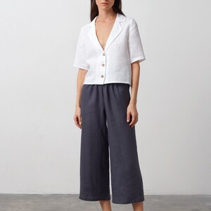 May include: A white linen shirt with a collar and short sleeves, paired with dark gray linen pants. The model is wearing black leather slip-on shoes.