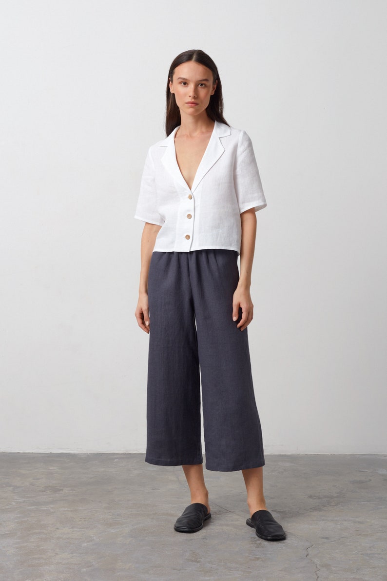 May include: A white linen shirt with a collar and short sleeves, paired with dark gray linen pants. The model is wearing black leather slip-on shoes.