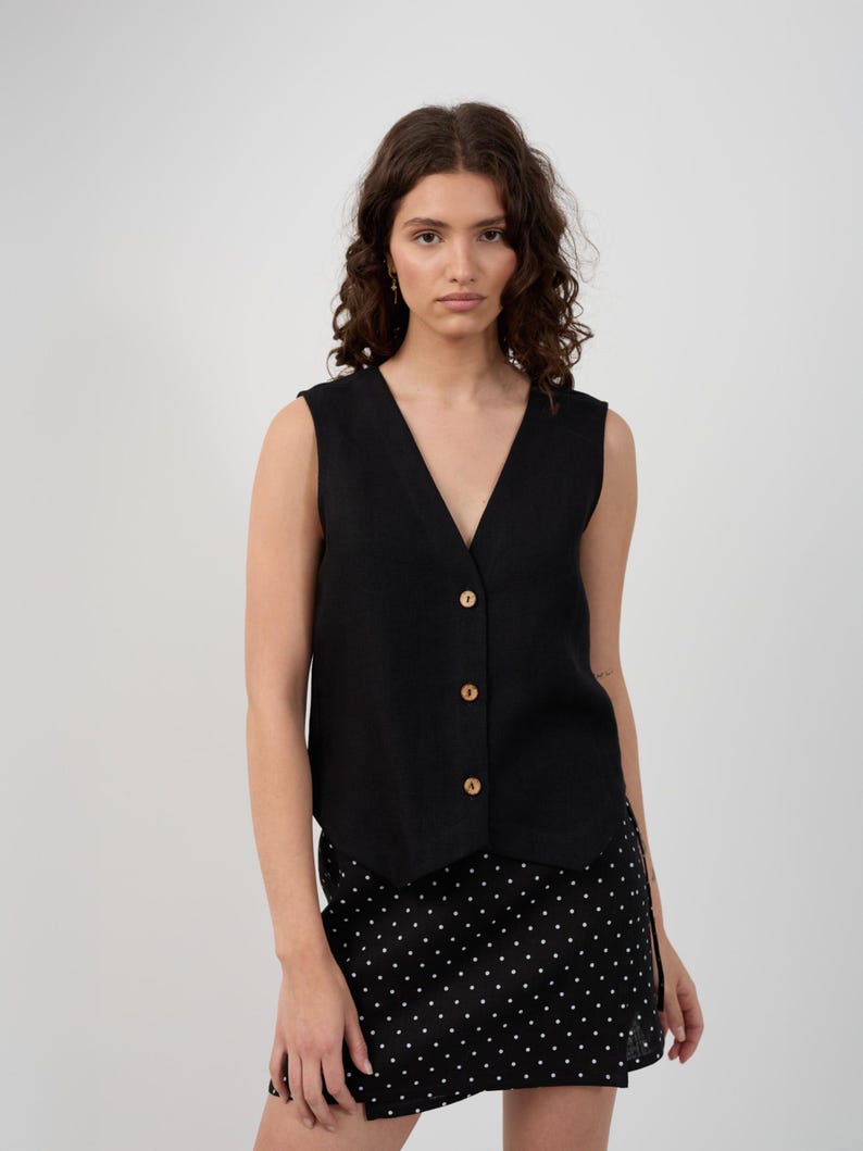 May include: A black sleeveless vest with a V-neckline and wooden buttons. The vest is paired with a black skirt with a white polka dot pattern. The outfit is simple and stylish.